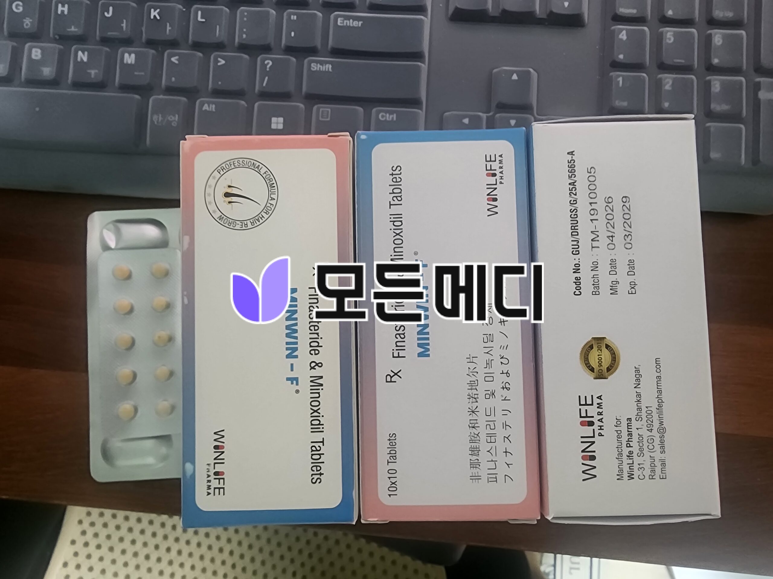 Row of medicine boxes on a desk in front of a computer keyboard, featuring blue, pink, and white packaging and a blister pack of pills on the left.