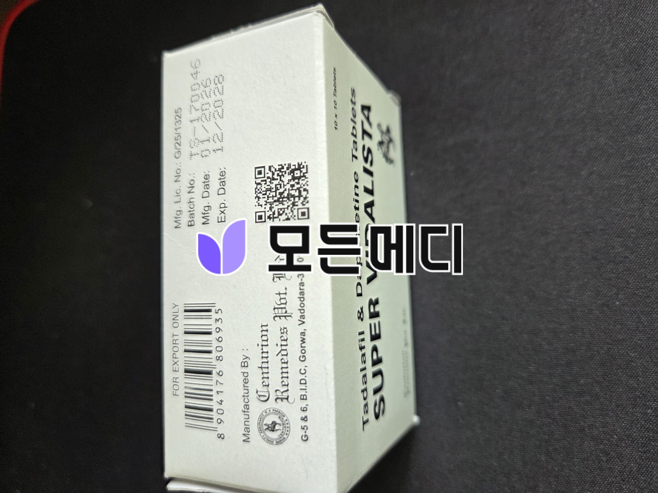 White rectangular medicine box angled on a dark surface, showing a barcode, printed text, a QR code, and a small purple logo partially visible on the side.