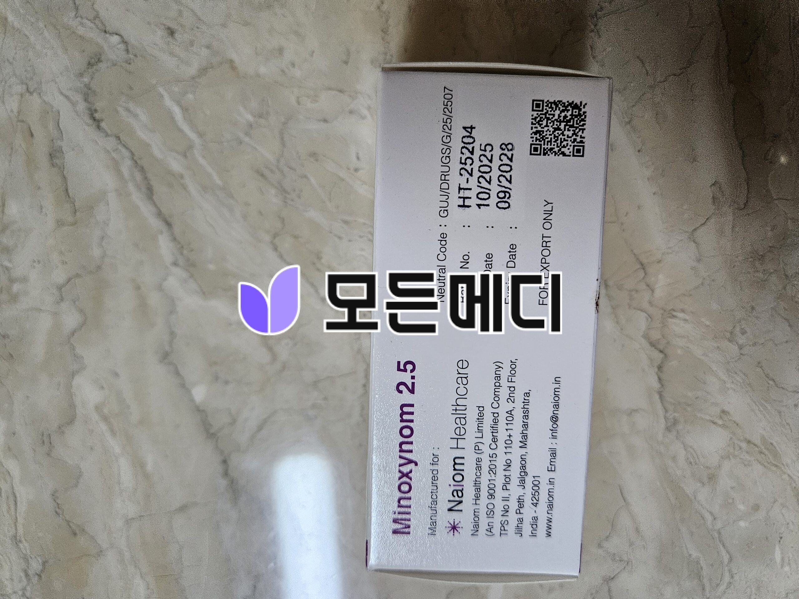 White rectangular medicine box on a marble surface, showing a purple logo and vertical black text with a QR code on the side.